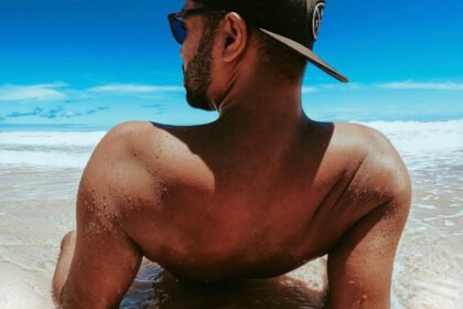 Muškarac sa kačketom i sunčanim naočarima sedi u plićaku na plaži.