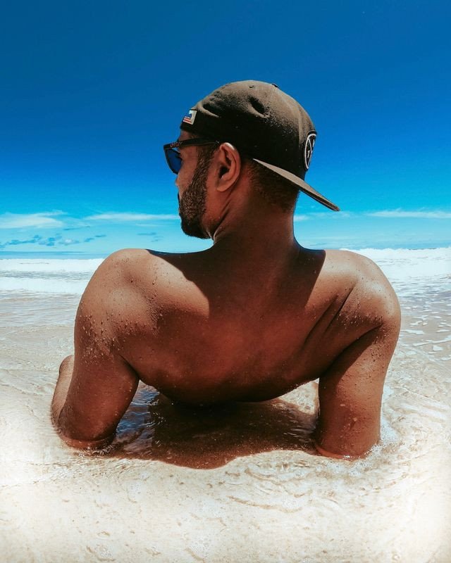 Muškarac sa kačketom i sunčanim naočarima sedi u plićaku na plaži.